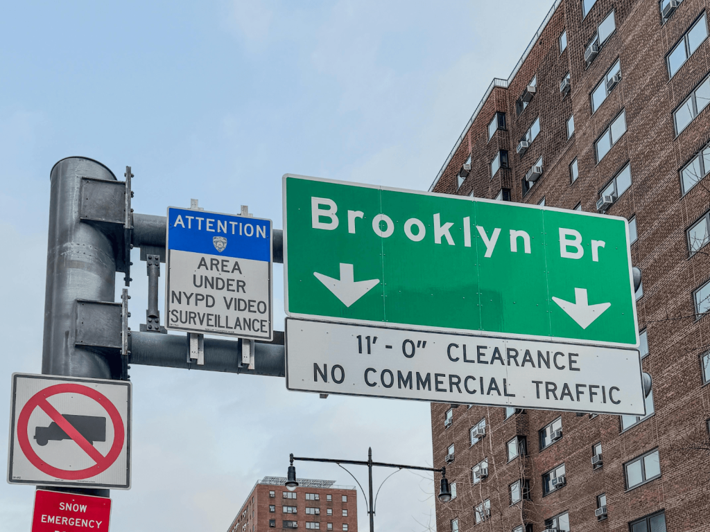 Green Brooklyn Bridge road sign surrounded by city buildings and street signs in New York City.