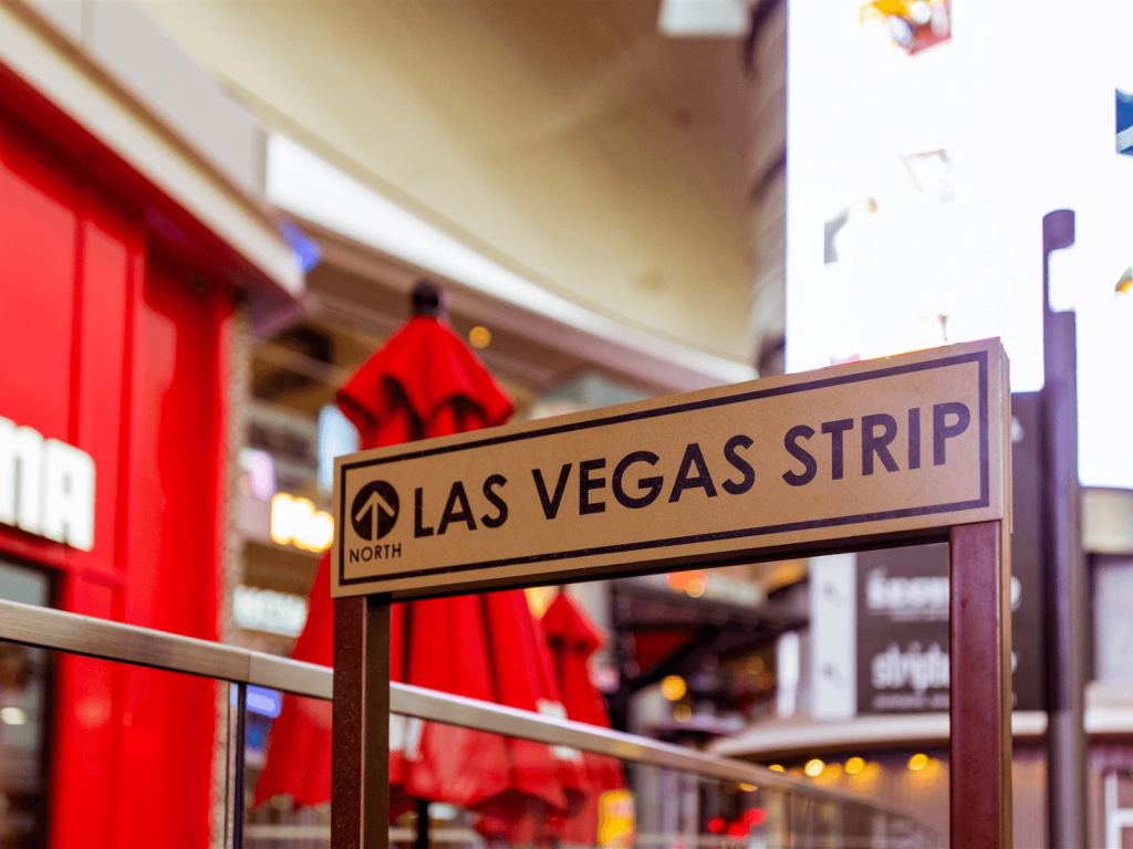 Sign pointing toward the Las Vegas Strip with bright signs and red umbrellas in the background, perfect for Las Vegas captions.