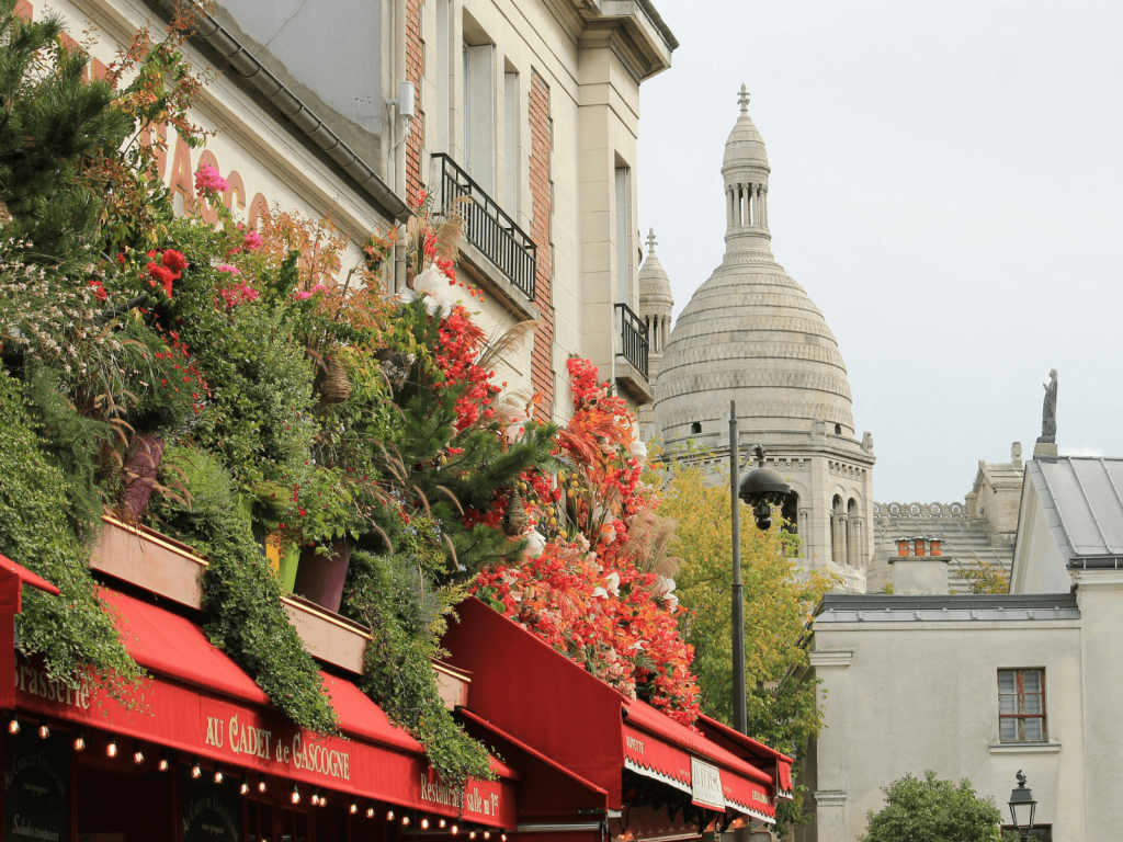 Colorful flowers and greenery covering a café in Montmartre with the dome of Sacré Coeur rising in the background, perfect for Paris Instagram captions.