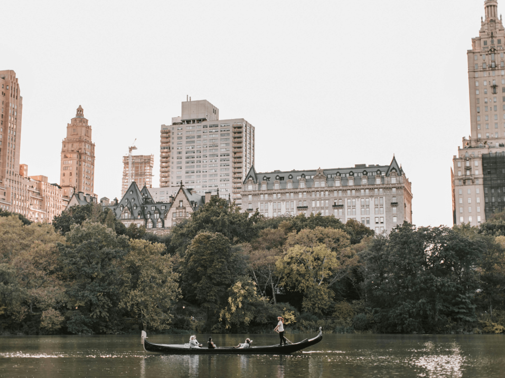 Gondola crossing the lake in Central Park with trees and classic Manhattan buildings in the background, perfect for Central Park captions.