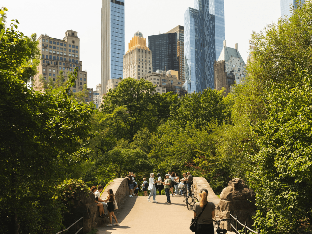 Visitors walking through a lush green section of Central Park with tall Manhattan skyscrapers rising above the trees.