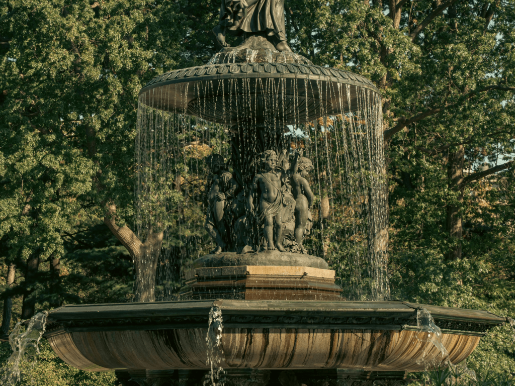 Ornate Bethesda Fountain surrounded by leafy trees in Central Park with water cascading around the sculptures.