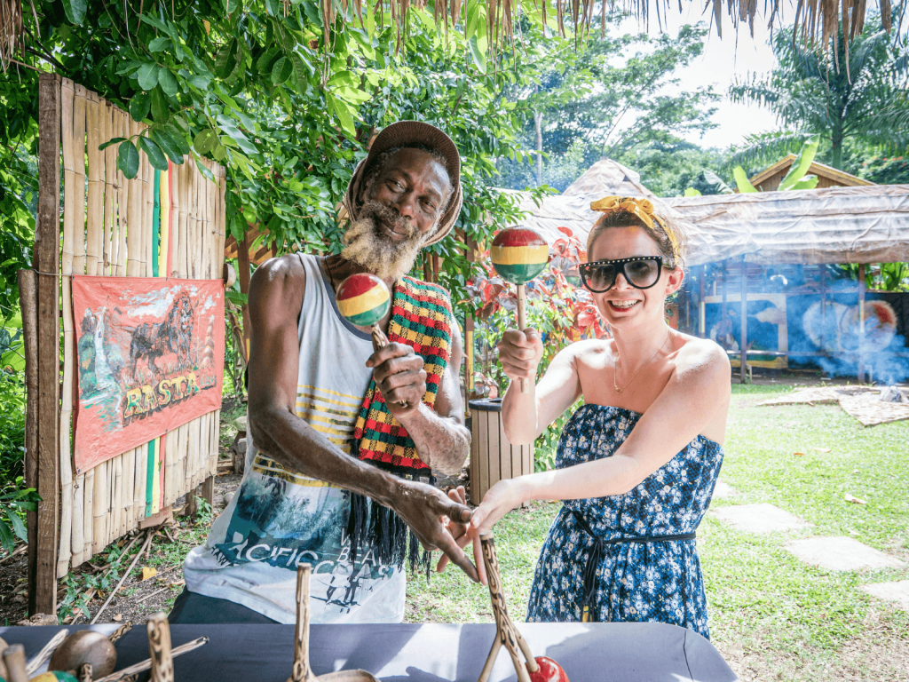 Woman smiling with a local musician while holding colorful maracas at a tropical outdoor setting in Jamaica.