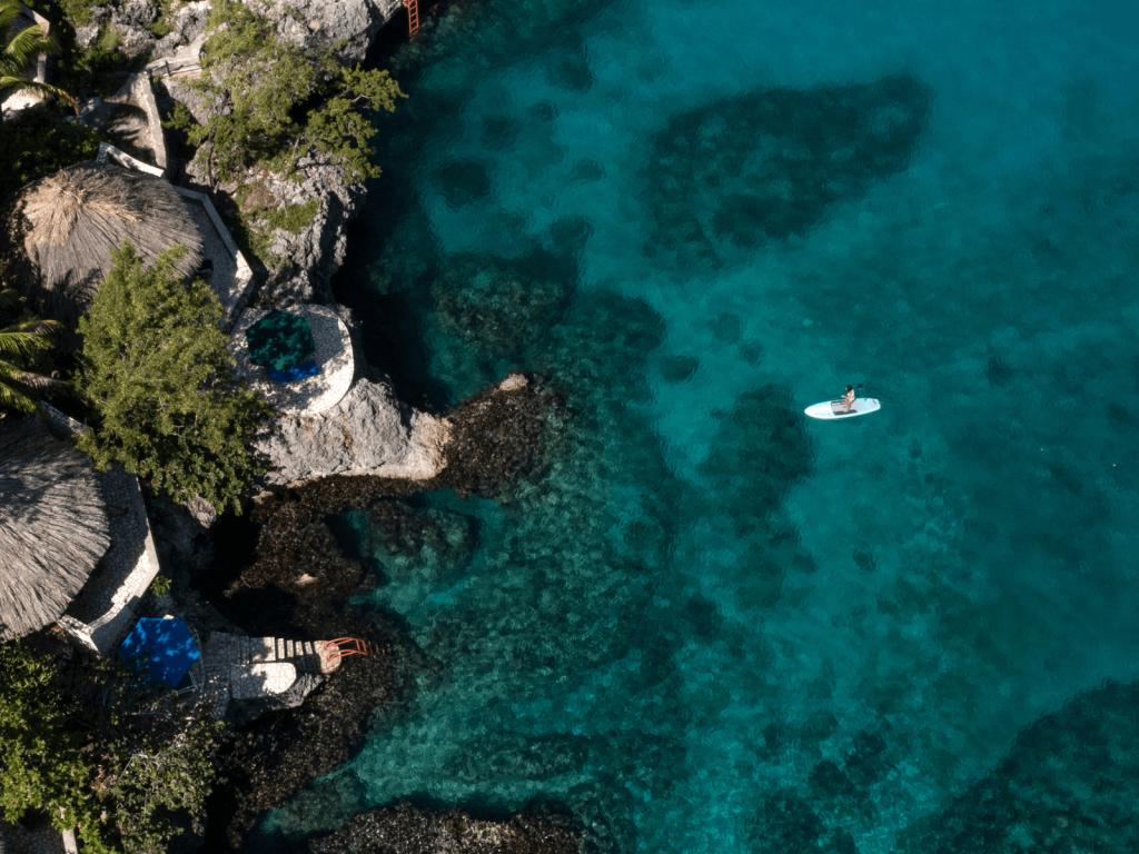Aerial view of a paddleboarder floating over clear turquoise Caribbean water beside a rocky resort coastline, perfect for Jamaica captions.