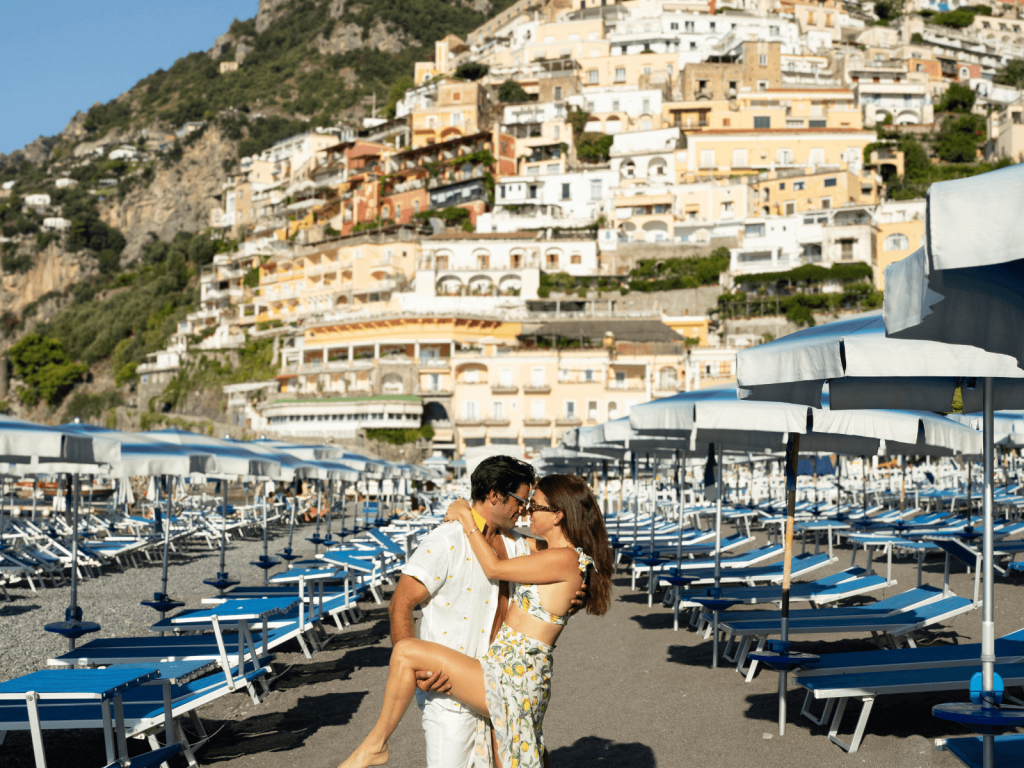 Couple embracing on a beach in Positano with rows of blue lounge chairs and colorful hillside buildings behind them.