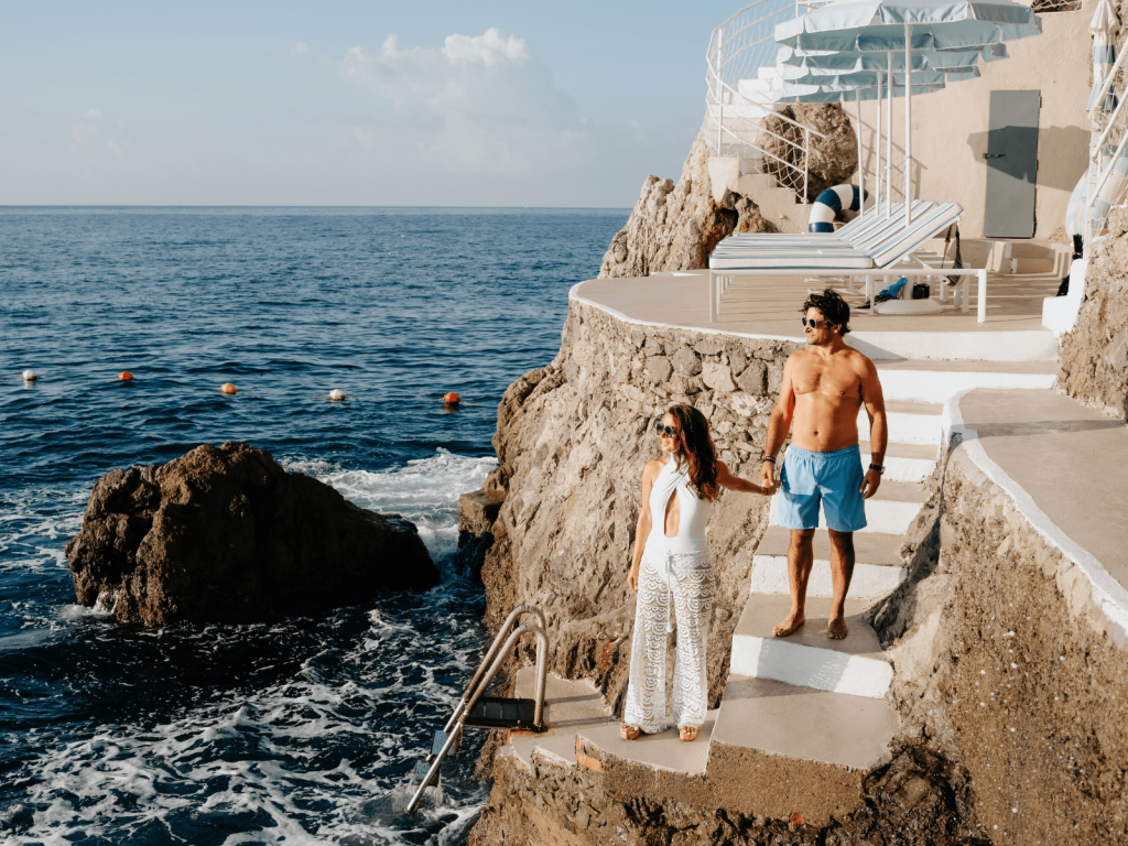 Couple holding hands on stone steps beside the Mediterranean Sea at a cliffside beach club in Italy.
