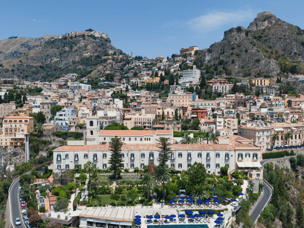 Panoramic view of Taormina, Sicily, with colorful buildings climbing the mountainside beneath a bright blue sky.
