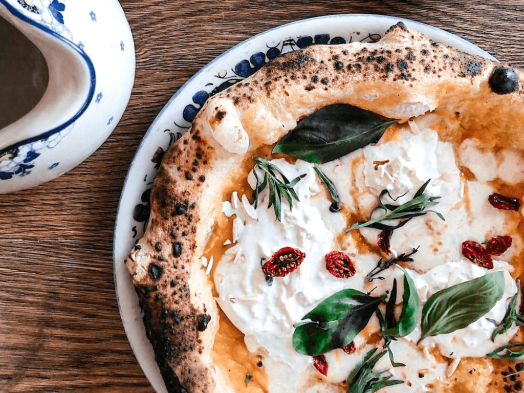 Close up of an Italian pizza topped with creamy cheese, basil, herbs, and sun dried tomatoes on a wooden table.