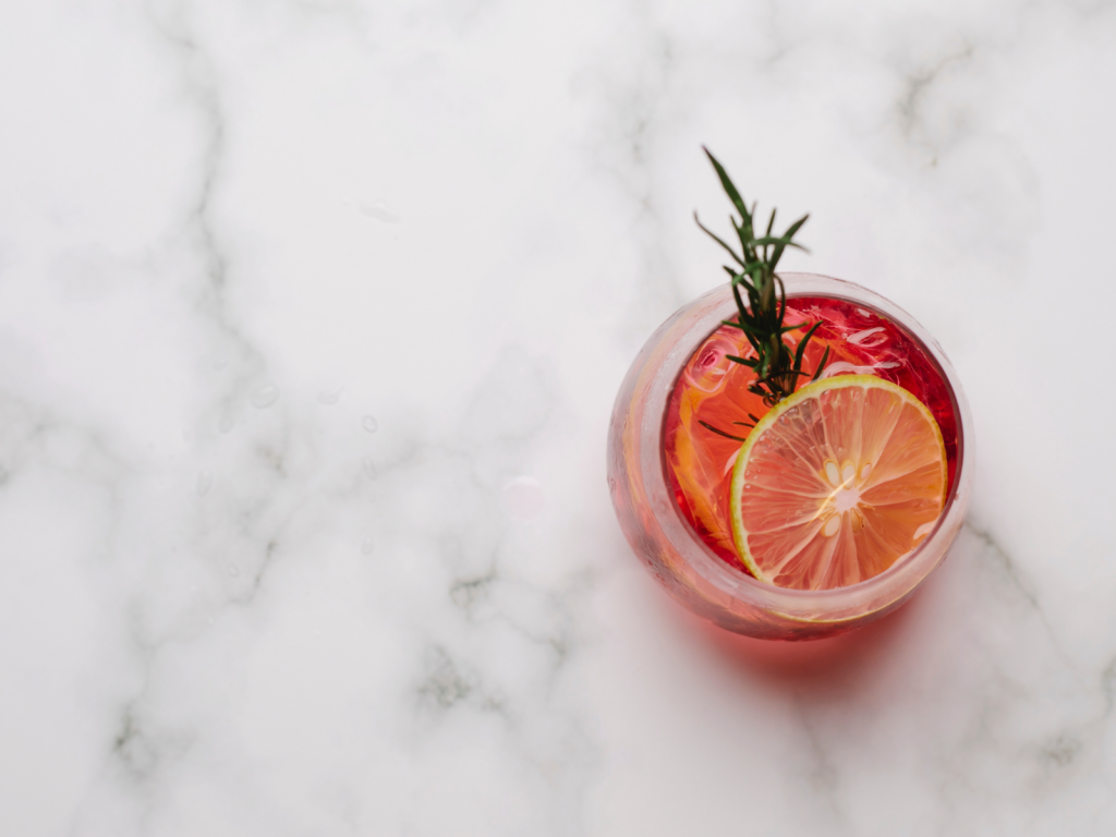 Pink cocktail with a lemon slice and rosemary garnish on a white marble surface, perfect for cocktail captions for Instagram.