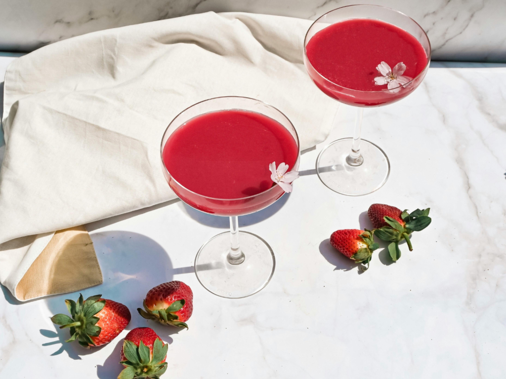 Two bright pink cocktails in coupe glasses garnished with small flowers, surrounded by fresh strawberries on a marble table.