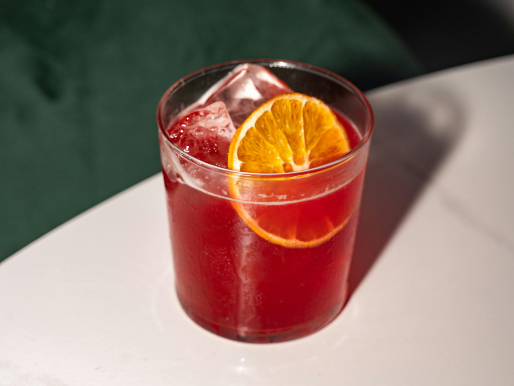 Bright red cocktail served over ice and garnished with a fresh orange slice on a white table.