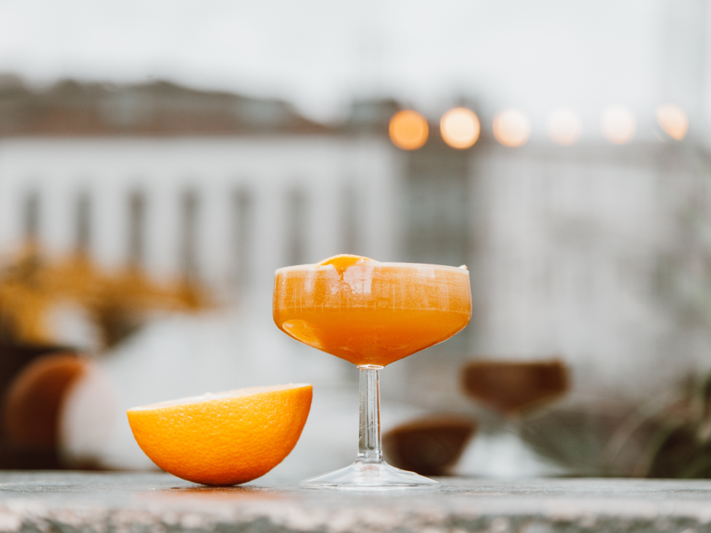 Orange cocktail in a coupe glass beside a fresh orange half, with a softly blurred outdoor background.
