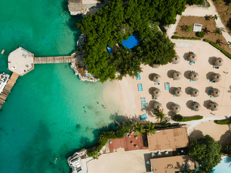 Aerial view of a Caribbean beach with turquoise water, a wooden pier, thatched umbrellas, and lounge chairs, perfect for Caribbean Instagram captions.