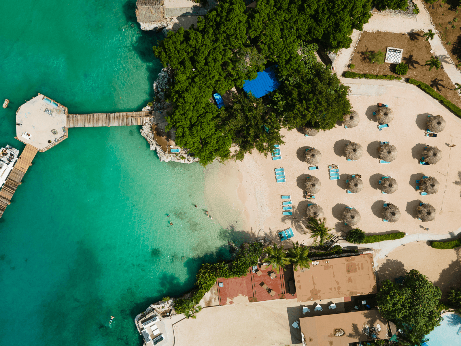 Aerial view of a Caribbean beach with turquoise water, a wooden pier, thatched umbrellas, and lounge chairs, perfect for Caribbean Instagram captions.