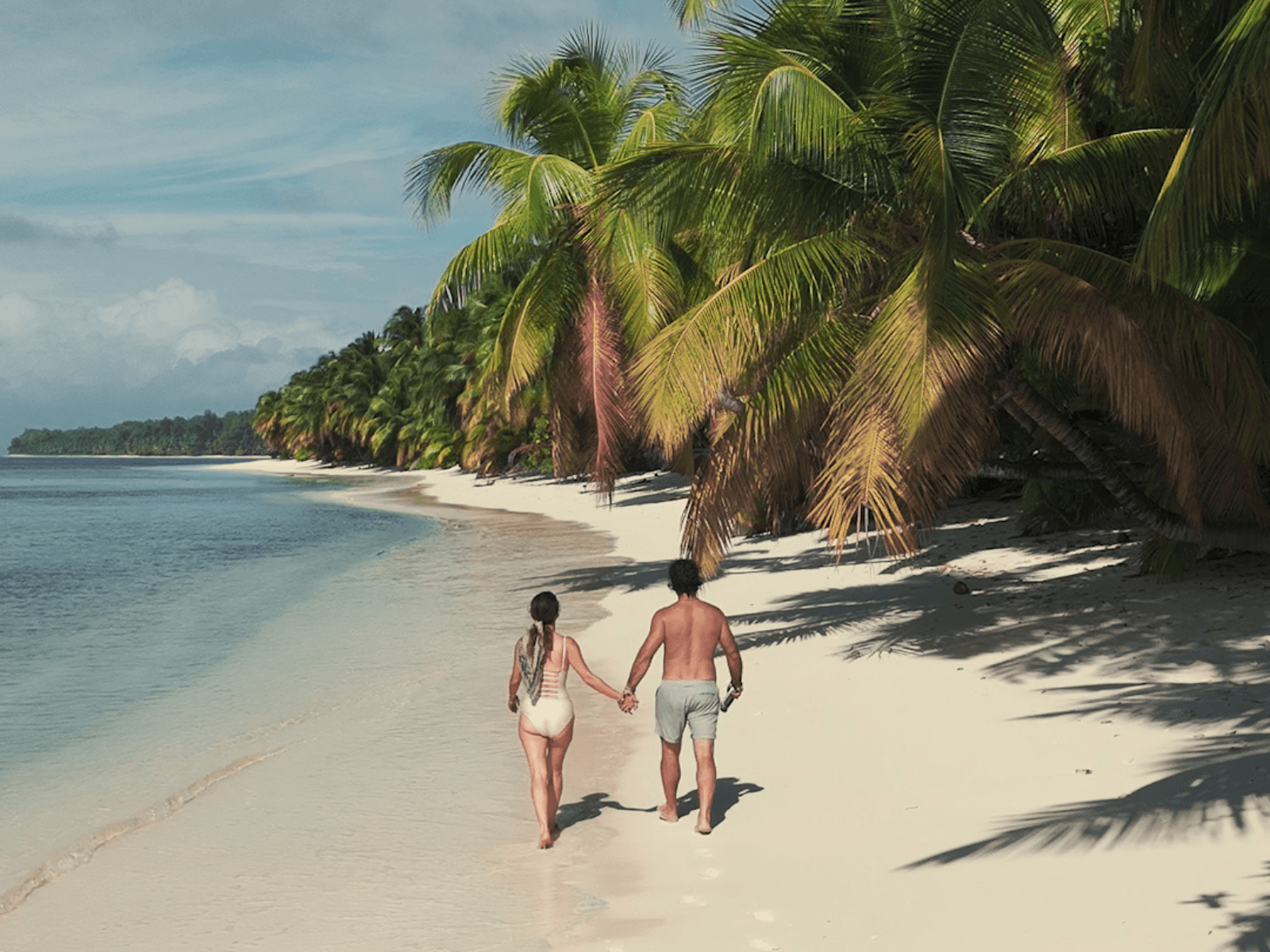 Couple holding hands while walking along a white sand beach lined with palm trees and turquoise water in the Seychelles, featured in a Seychelles itinerary.