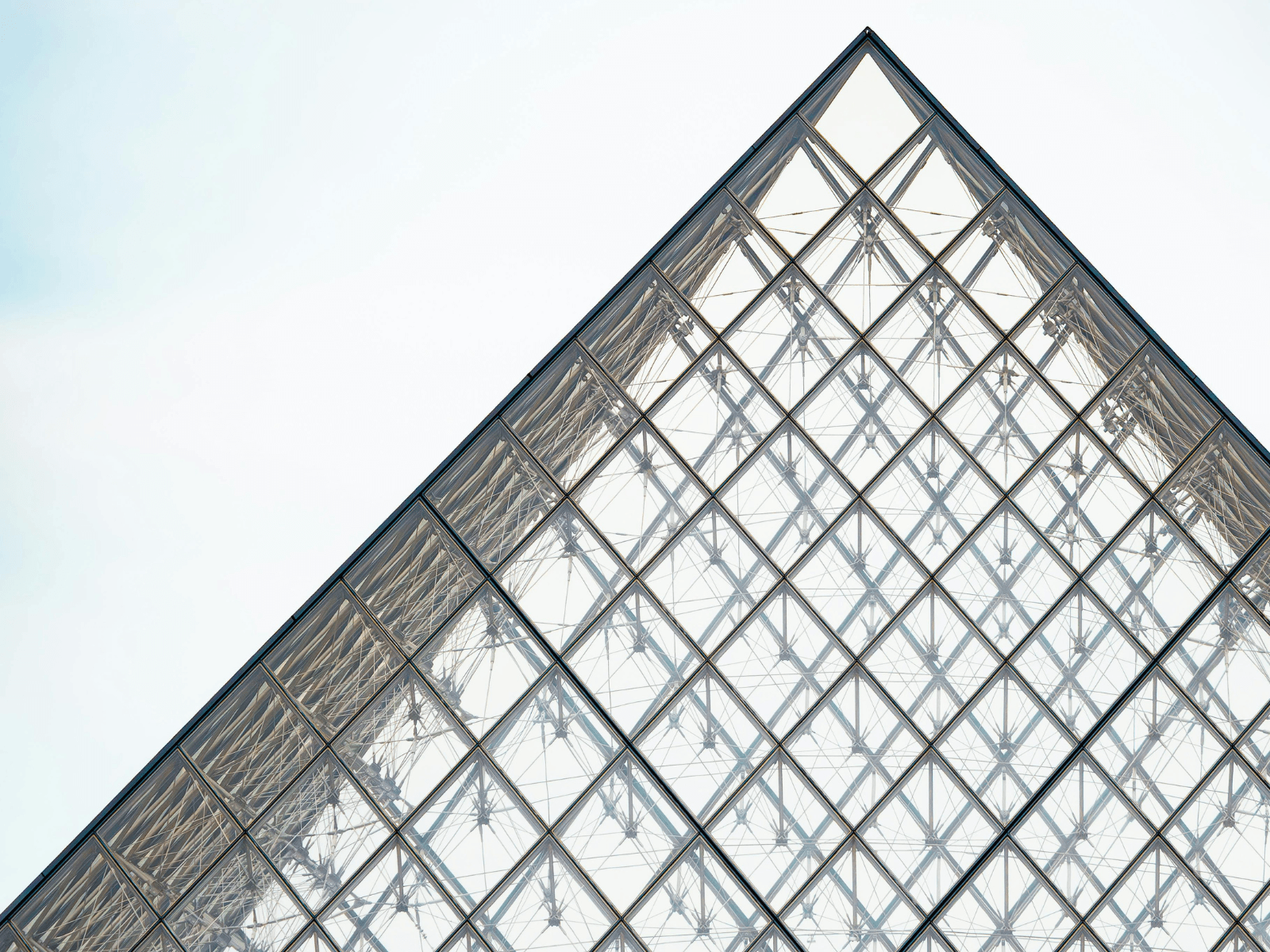 Close up view of the Louvre Pyramid in Paris, showing its geometric glass panels and metal framework against a bright sky, perfect for Louvre captions.