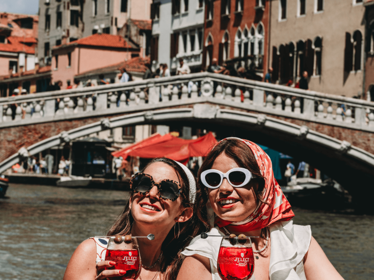 Two women wearing sunglasses and headscarves enjoy red spritz cocktails beside a canal and bridge in Venice, Italy, during a one day in Venice trip.