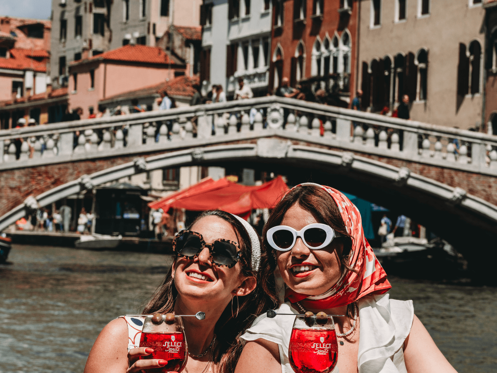Two women wearing sunglasses and headscarves enjoy red spritz cocktails beside a canal and bridge in Venice, Italy, during a one day in Venice trip.