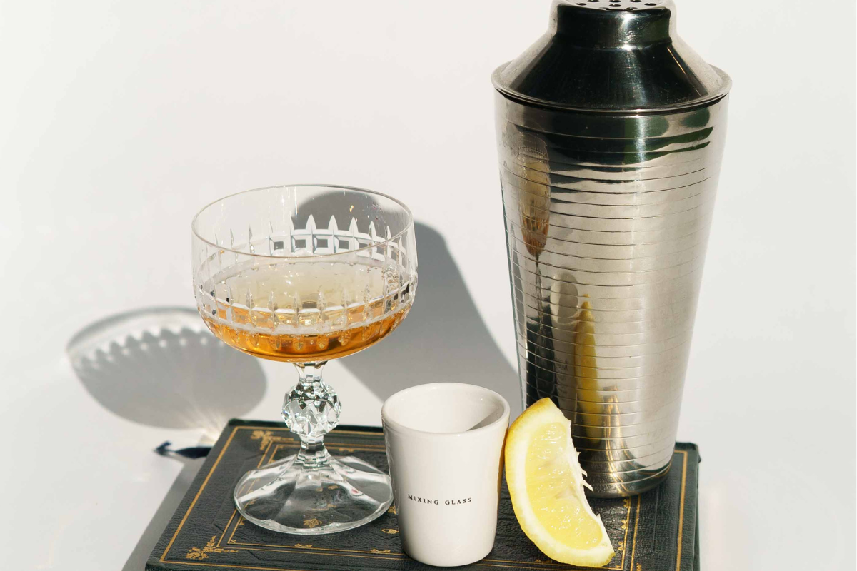 Home bar essentials including a cocktail shaker, coupe glass with a cocktail, mixing glass, and fresh lemon wedge.