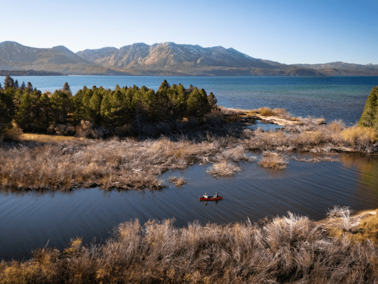 Aerial view of two people canoeing through calm water surrounded by trees and brush, with Lake Tahoe and mountains in the background, for a post about Lake Tahoe captions.