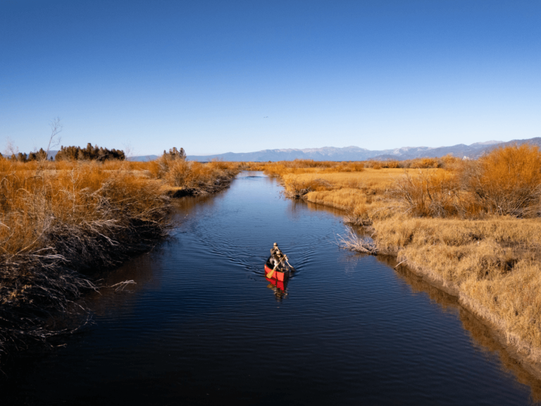 Two people canoeing along a calm river surrounded by golden grasses with snowcapped mountains near Lake Tahoe for a Lake Tahoe itinerary.
