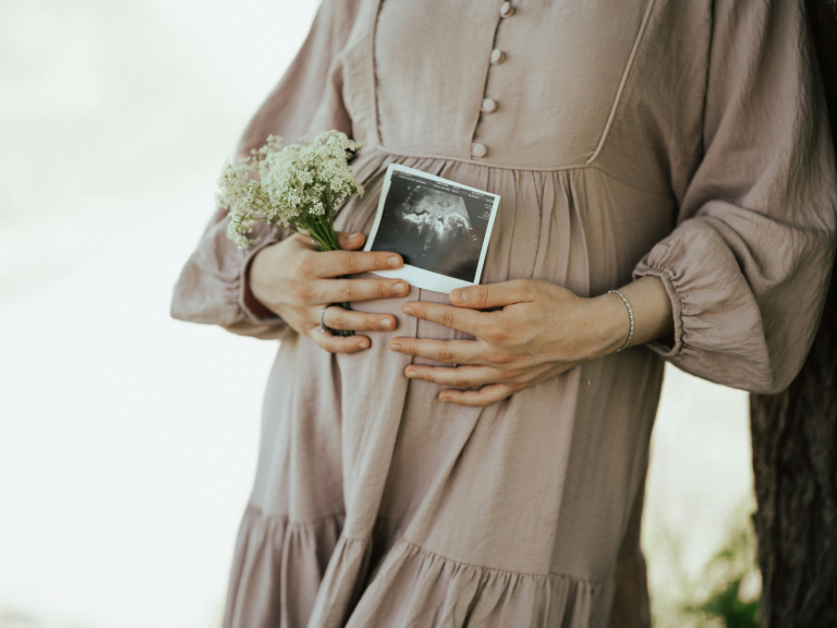 Pregnant woman holding an ultrasound photo and flowers over her baby bump for a collection of pregnancy announcement captions.