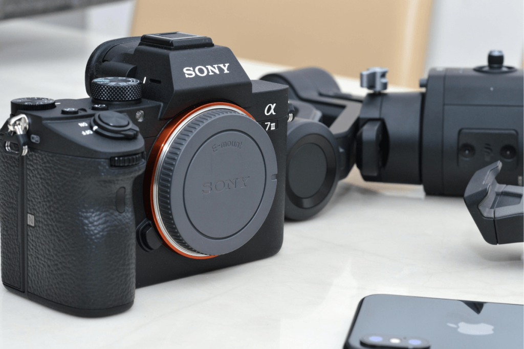 Sony Alpha mirrorless camera displayed with additional camera gear, including a camera stabilizer and smartphone, on a white tabletop.