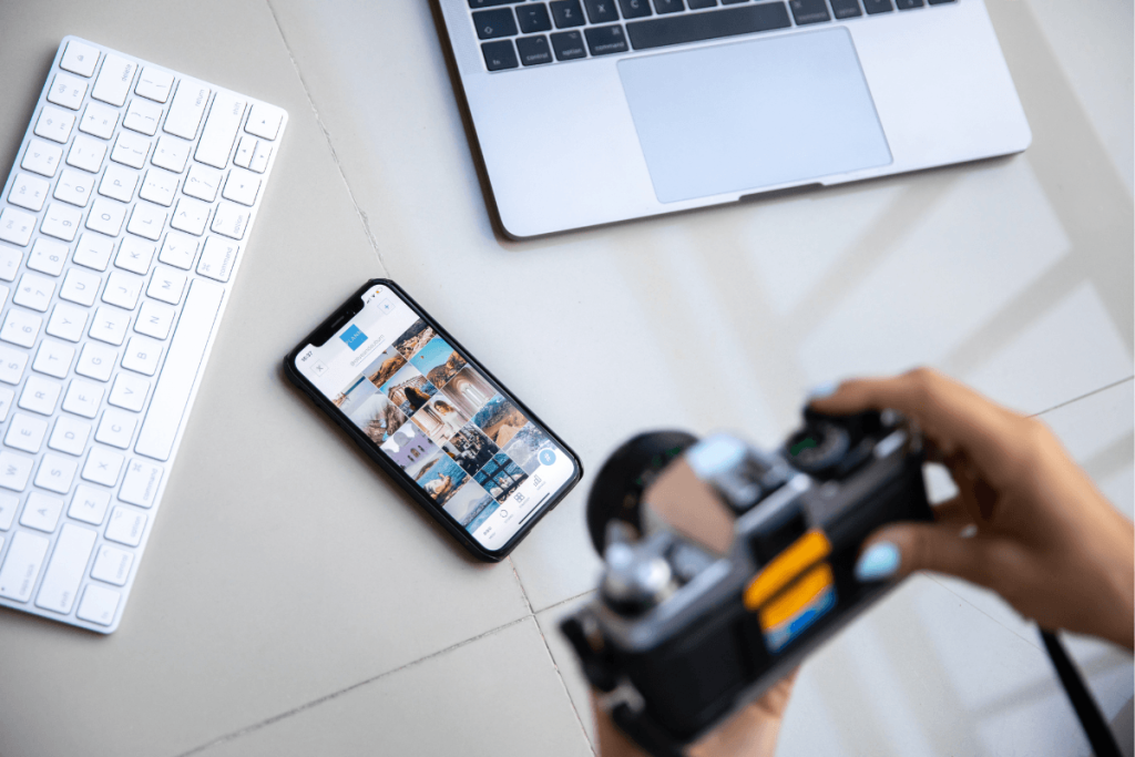 Photographer holding a camera beside a laptop, keyboard, and smartphone displaying a gallery of travel photographs.