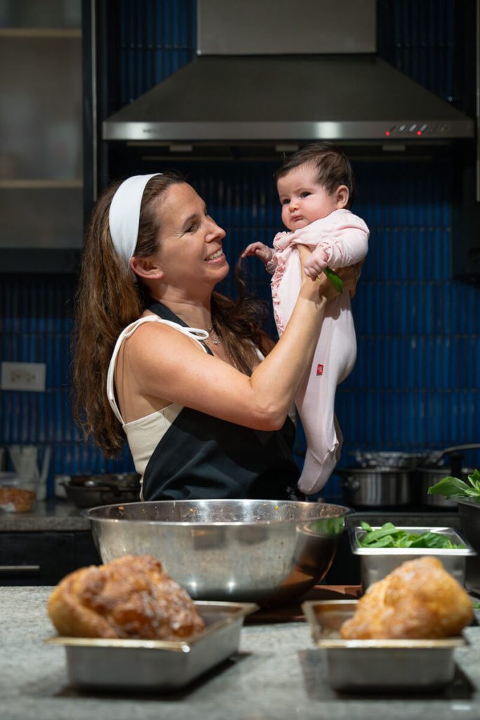 A mother smiling while holding her baby in a cozy kitchen, surrounded by bowls and fresh ingredients — capturing sweet everyday joy.