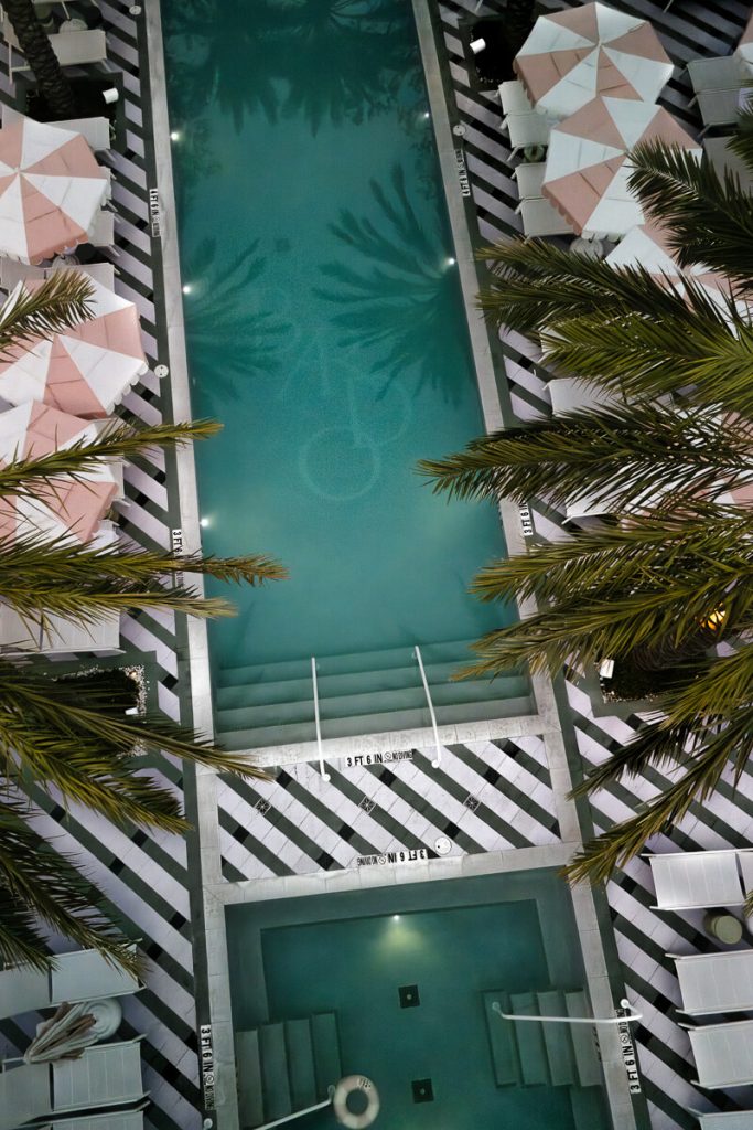Aerial view of a chic hotel pool lined with pink and white umbrellas, palm trees, and striped tile patterns — the perfect relaxing spot for 2 days in Savannah, Georgia.