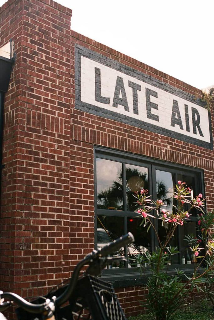 Exterior of a red brick building with “Late Air” painted above the windows, framed by blooming flowers and a vintage bike.