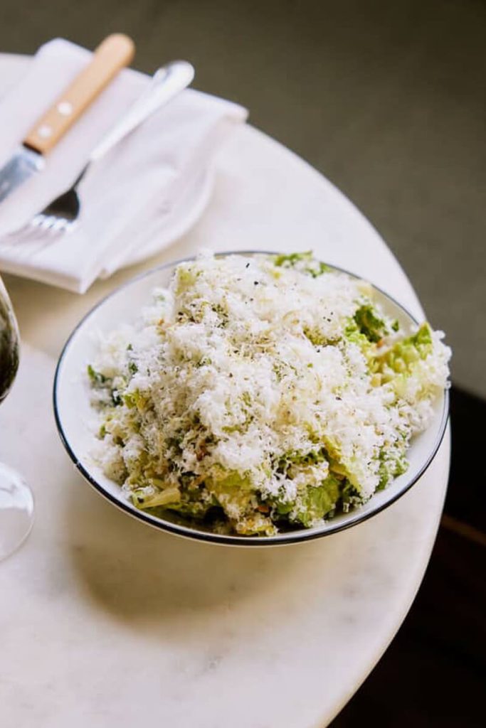 A bowl of Caesar salad topped with a generous layer of grated cheese on a marble table, paired with wine and simple dining setup.