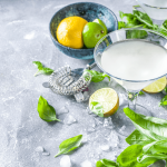 A refreshing cucumber basil gimlet served in a cocktail glass, garnished with fresh basil and surrounded by limes, lemons, and scattered basil leaves on a gray stone surface.