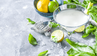 A refreshing cucumber basil gimlet served in a cocktail glass, garnished with fresh basil and surrounded by limes, lemons, and scattered basil leaves on a gray stone surface.