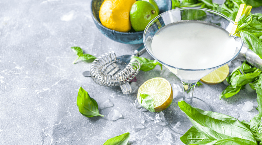 A refreshing cucumber basil gimlet served in a cocktail glass, garnished with fresh basil and surrounded by limes, lemons, and scattered basil leaves on a gray stone surface.