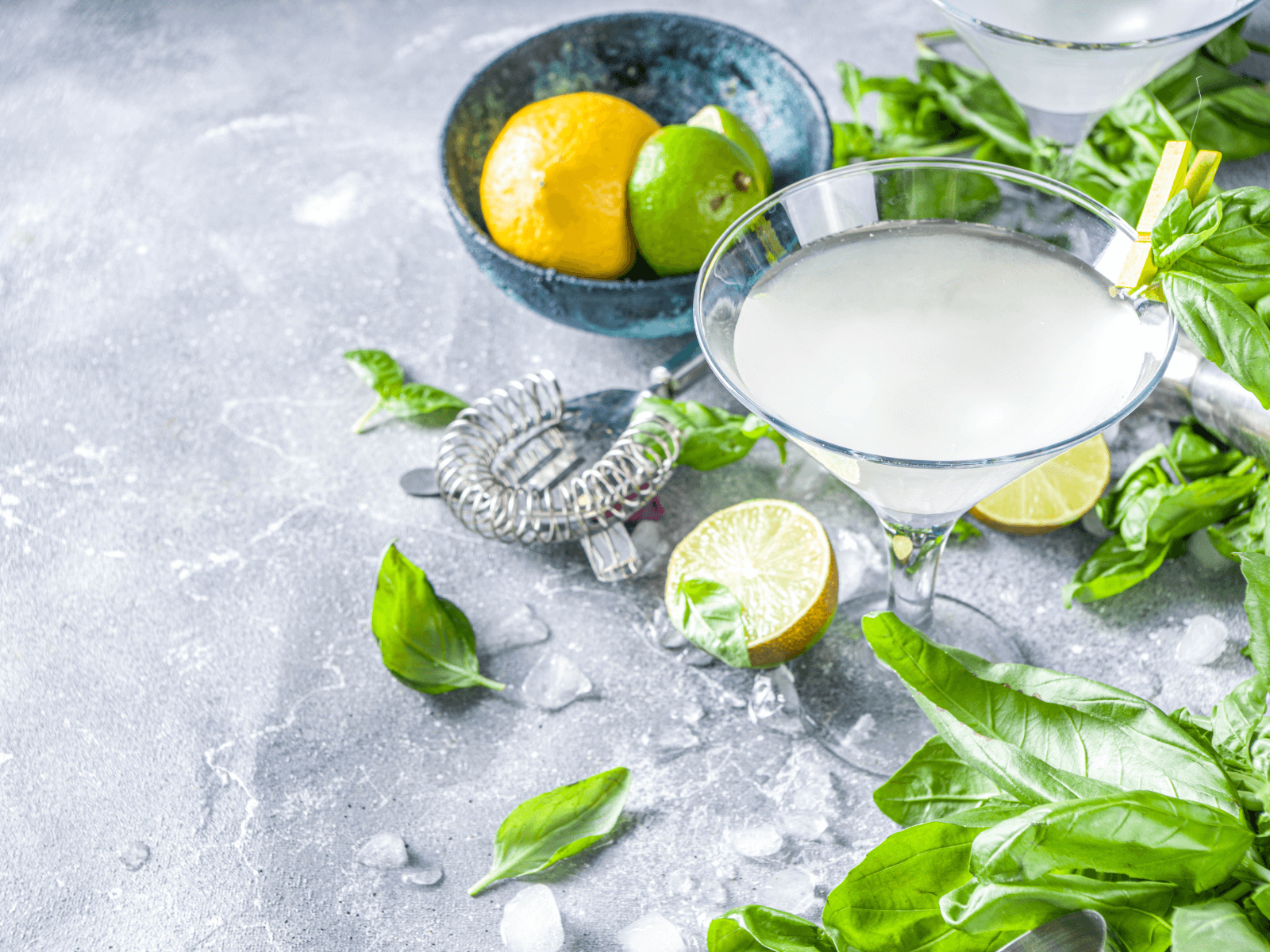A refreshing cucumber basil gimlet served in a cocktail glass, garnished with fresh basil and surrounded by limes, lemons, and scattered basil leaves on a gray stone surface.