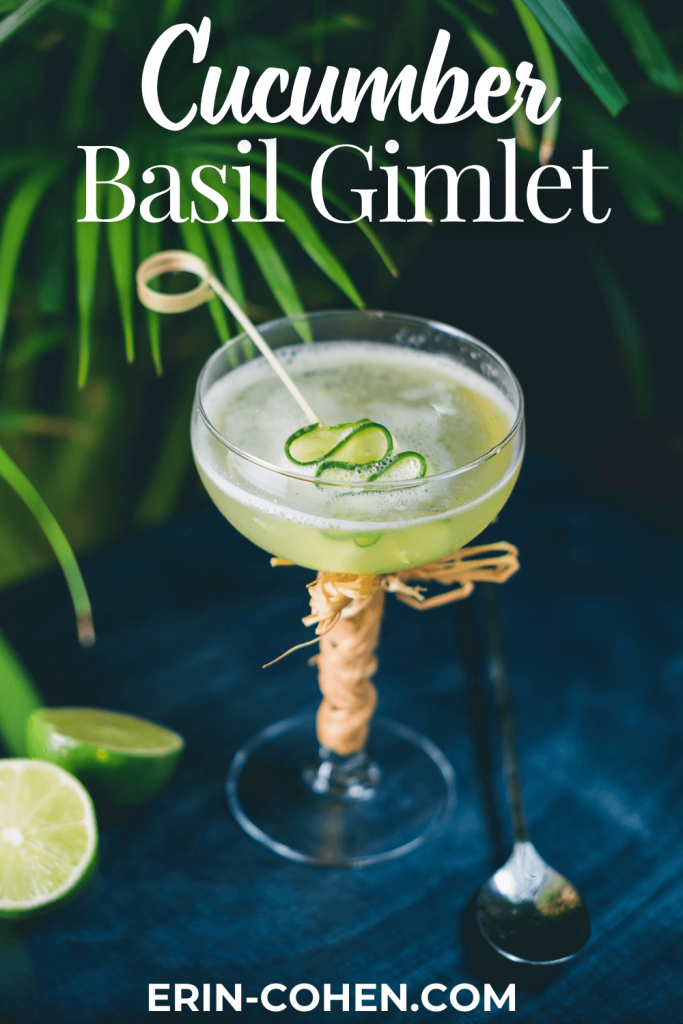 A fresh cucumber basil gimlet in a coupe glass, garnished with cucumber ribbons and a bamboo skewer, sits on a dark table next to lime wedges, with tropical green leaves in the background and “Cucumber Basil Gimlet” text above.