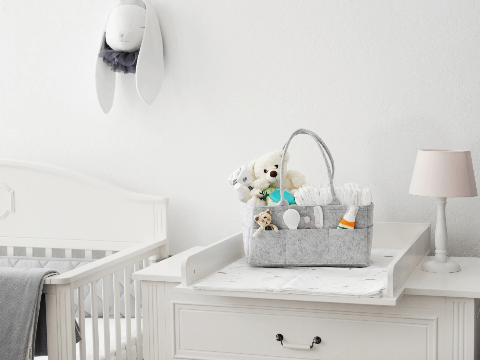 Soft neutral nursery with a diaper changing station topped with a grey organizer basket filled with stuffed animals, baby lotion, diapers, and baby care items. Showcasing practical newborn diaper bag essentials in a cozy and minimalist setup.