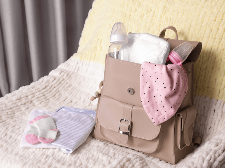 A tan diaper bag sits open on a soft white and yellow blanket, packed with baby travel essentials including diapers, baby bottles, and a pink polka-dot bib. Next to it are folded baby clothes and tiny socks. This visual represents a well-prepped baby travel packing list, offering a glimpse of what parents might bring for a trip with their little one.