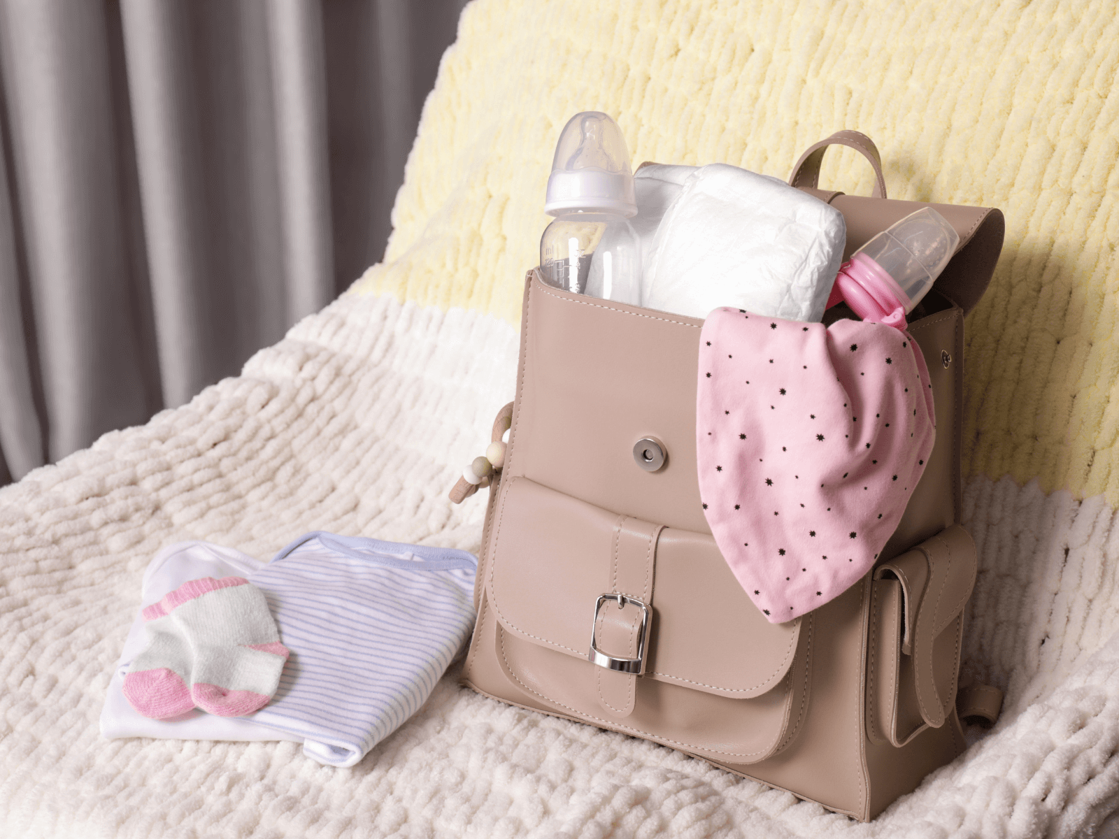 A tan diaper bag sits open on a soft white and yellow blanket, packed with baby travel essentials including diapers, baby bottles, and a pink polka-dot bib. Next to it are folded baby clothes and tiny socks. This visual represents a well-prepped baby travel packing list, offering a glimpse of what parents might bring for a trip with their little one.