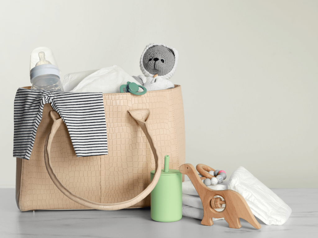 Tan diaper bag filled with baby must-haves including a striped onesie, baby bottle, diapers, pacifier, stuffed animal, and wooden toys—perfect example of Newborn Diaper Bag Essentials.