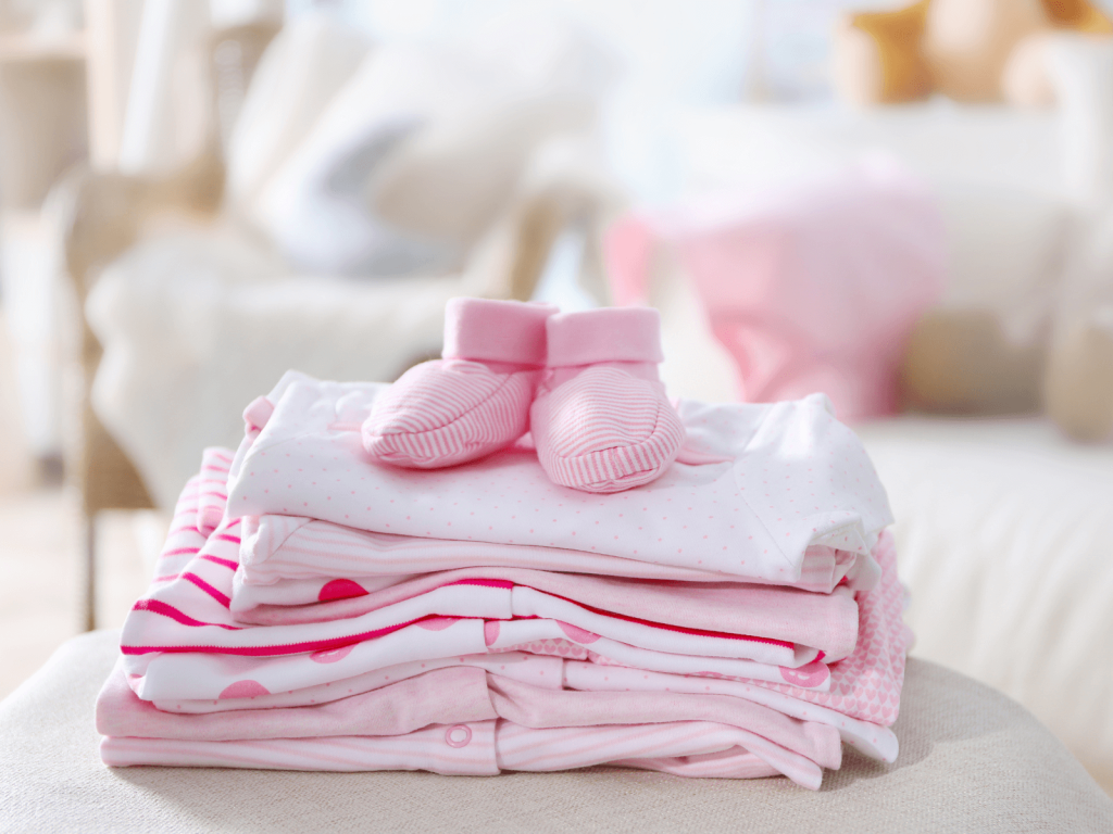 Neatly folded pink baby clothes with soft booties stacked on a chair in a cozy nursery—organized and ready for a newborn.