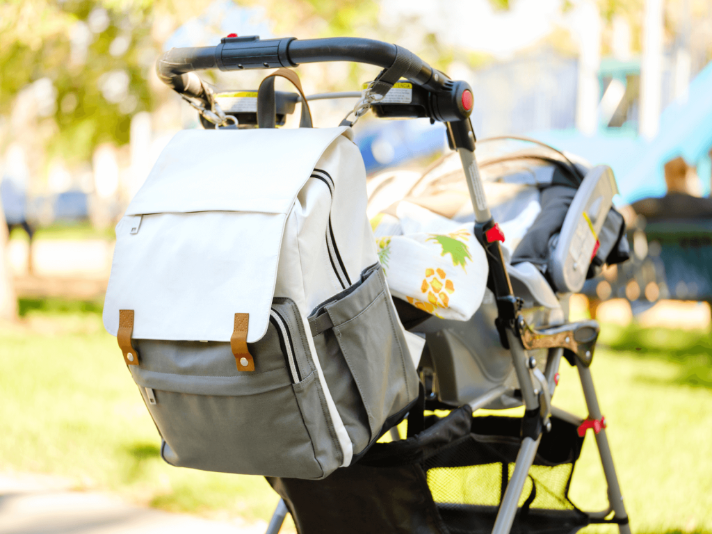 Modern white and gray diaper bag hanging on a stroller in a sunny park—packed and ready with all the Newborn Diaper Bag Essentials for an outing.