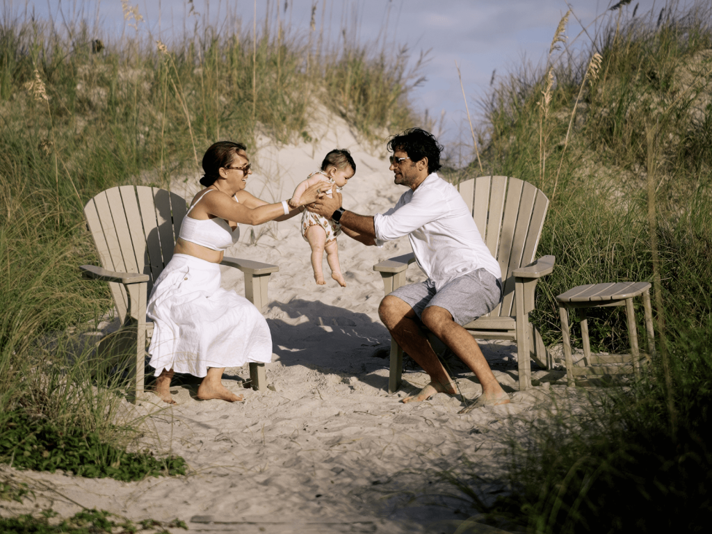A joyful moment between a couple and their baby on a sandy path through the dunes. They sit on beach chairs, facing each other as the dad playfully lifts the baby from mom’s hands. Tall beach grass and blue skies frame the scene.