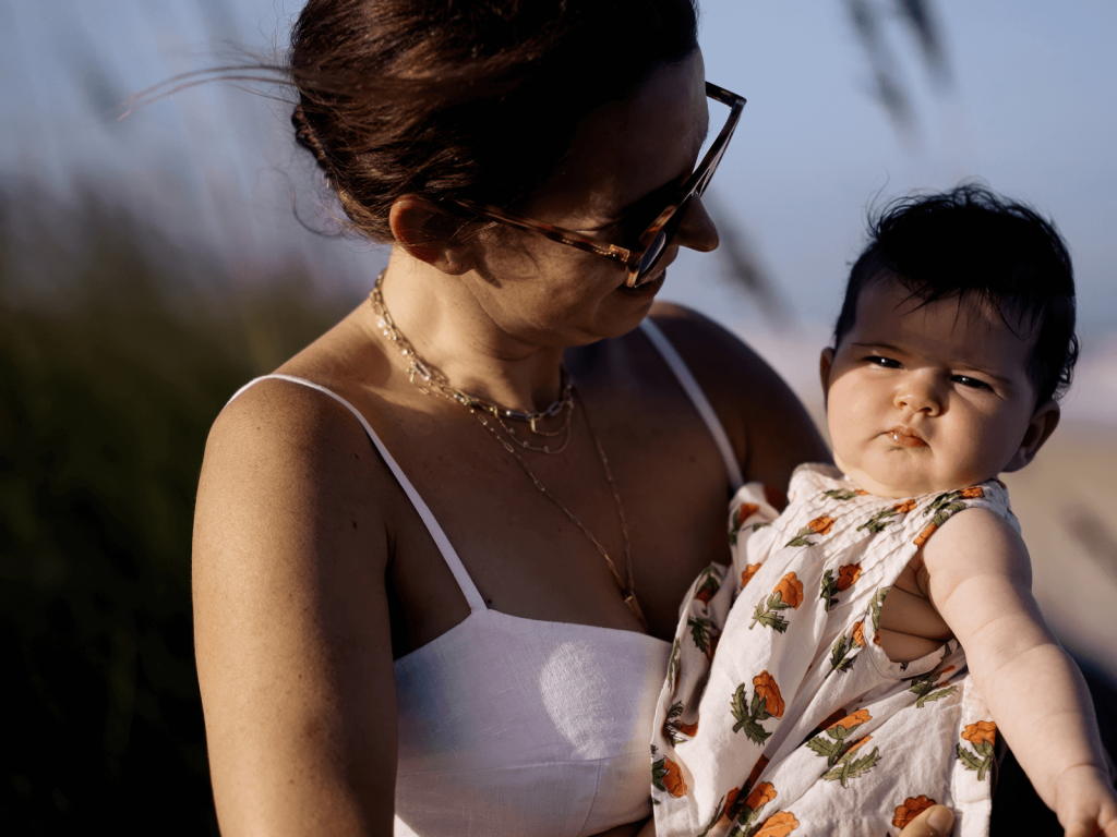 A woman in a white dress holds a baby wearing a floral sundress. The woman smiles down at the baby while the baby looks directly at the camera with a serious expression. Sunlight highlights their faces, with beach grass blurred in the background.