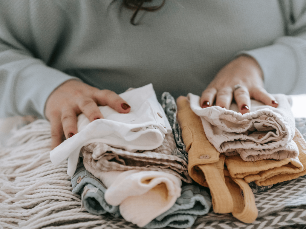Person organizing folded neutral colored baby clothes on a bed, including onesies, pants, and other newborn diaper bag essentials.