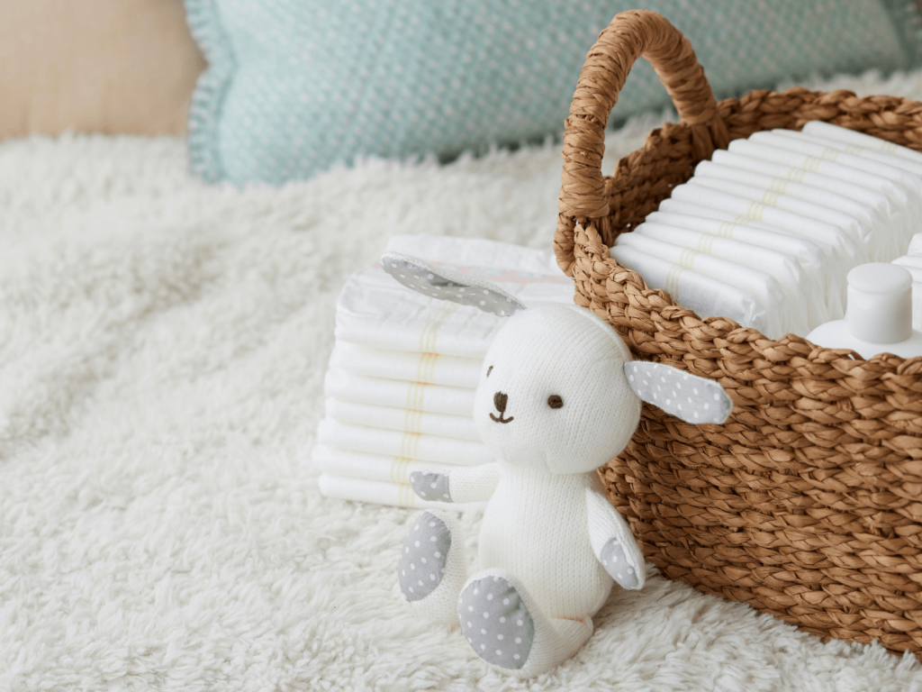 Woven basket filled with disposable diapers beside a soft stuffed lamb and additional diapers on a nursery bed.