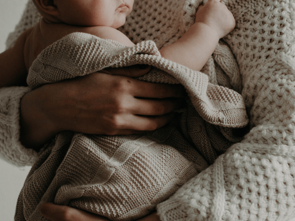 Parent holding a newborn baby wrapped in a soft knitted blanket against their chest.