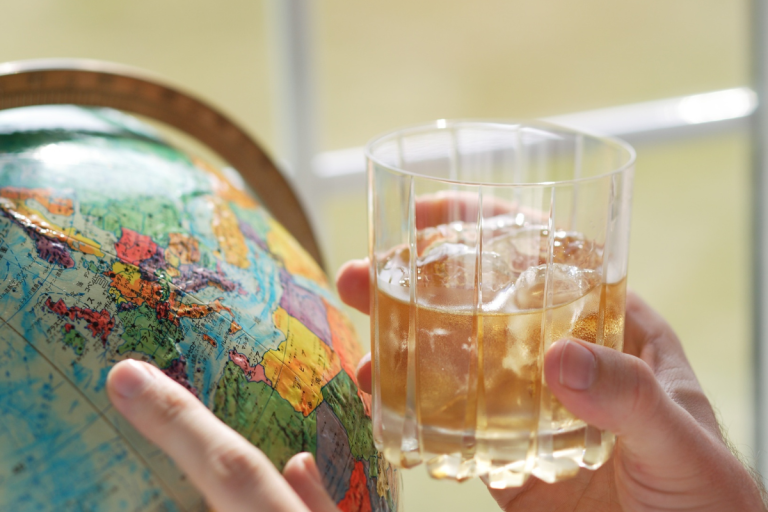 Person holding a whiskey cocktail with ice beside a globe for a guide to cocktails around the world.
