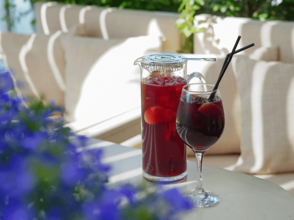 Glass and pitcher of red sangria with fresh fruit and black straws on a sunny outdoor table.